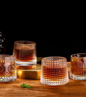Multiple styled whiskey tumblers from a premium OEM whiskey glass supplier, showcasing ribbed, waffle, and vintage embossed textures on a wooden table, ideal for bulk custom hospitality drinkware manufacturing.