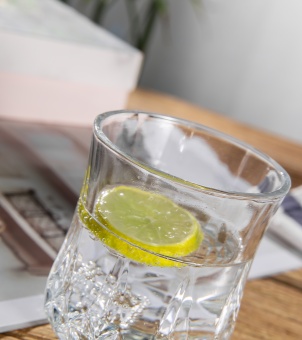 Close-up shot of a cafe-style crystal cut glass cup filled with clear water and a fresh lemon slice, set at an angle on a wooden table with blurred magazine background.