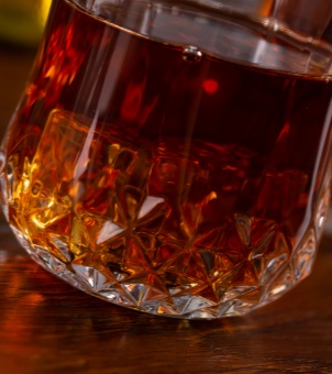 Close-up view of a heavy base commercial-grade whiskey glass featuring an intricate crystal cut diamond pattern filled with dark amber liquor on a wooden counter.