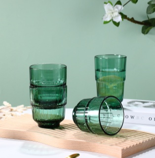Green tinted glass tumblers on wooden surface Several green glasses are placed on a wooden surface, with two glasses stacked together. In the background, there is a book and a flower.
