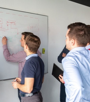 A group of male engineers and designers collaborating during a whiteboard session. One team member draws a system architecture workflow with red and green markers, while others look on attentively, holding a tablet and analyzing the diagram.