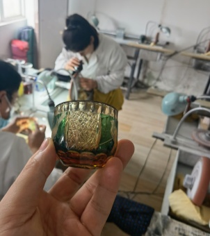 The image shows a glass with an engraved pattern on its surface, with a workshop where glass surface processing is done in the background.