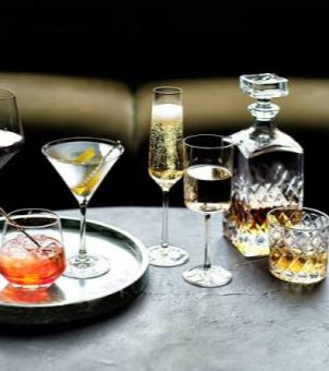 A meticulously arranged close-up photograph showcasing the product line of a high-end crystal glassware and home living brand. On a vintage round table with a natural, dark gray, rough texture, several exquisite high-end glassware pieces and prepared drinks are elegantly displayed. On the left is a round, silver, mirrored metal tray. To the left of the tray, in the foreground, sits a stout, baseless rock glass filled with a red nigroni or chilled cocktail, accompanied by a metal stirrer. To the right of the tray stands a classic inverted triangular martini glass, its clear liquid garnished with a sprig of lemon peel. Behind it, in the center of the table, stand two slender stemmed glasses side-by-side: on the left, a tall, cylindrical champagne flute filled with golden, finely bubbly champagne; on the right, a sleek, straight-sided white wine glass filled about halfway with a clear, light-colored fruit wine. On the right side of the image is a luxurious crystal engraving series: in the background is a square-faceted whiskey decanter with a square glass stopper and the lower half of the bottle engraved with fine diamond facets, containing amber-colored spirit; in the foreground is a matching classic whiskey glass with the same dense diamond-cut pattern, the liquid shimmering with a golden light under the refraction. The background is a dark-toned modern urban bar or subtly luxurious living room environment, with a curved-back sofa with a dark gold or leather sheen faintly visible in the background. The entire image is meticulously crafted with light and shadow, possessing excellent clarity, perfectly showcasing the luxurious texture of high-quality lead-free crystal glassware and the subtly luxurious atmosphere of modern refined living.