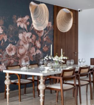 This is a real-life photo taken inside a high-end business hotel banquet hall or a modern, light luxury restaurant, showcasing professional tableware and interior design. The file name is Hospitality & Foodservice Dining room.jpg. The main subject of the image is a large, rectangular, pure white marble banquet table with legs made of white solid wood featuring multi-layered, vintage gourd-shaped carvings. The table surface is neatly arranged with a complete set of transparent crystal glassware for multiple seats, including stemmed red and white wine glasses placed one in front of the other, their surfaces reflecting a delicate white light under the lamps. A delicate bouquet of white and green flowers blooms in the center of the table, flanked by two slender, classic white candles. Surrounding the table are several exquisitely crafted modern solid wood armchairs, featuring walnut-colored wooden frames, off-white soft cushions, and vintage woven rattan mesh backrests. On the left side of the background is a striking dark blue-black art wallpaper wall, adorned with large, layered patterns of light pink and brown classic floral designs (such as lilies and roses); on the right, a vertically assembled dark solid wood wainscoting panel hangs. Above the dining table, two large, light-colored, double-layered, wavy, spiral rattan pendant lights, their intricate lines emitting a soft, translucent glow. The entire image creates a rich sense of space and warm lighting, seamlessly integrating highly transparent glassware into a modern, upscale banquet space imbued with a natural and vintage artistic ambiance.