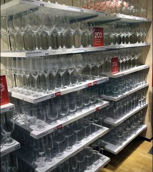 This is a real-life photo taken inside a modern department store or large chain supermarket, showing a sample display of glassware shelves. The file name is Retail & Supermarket Chains.jpg. The main subject of the image is a multi-tiered, pure white, modern supermarket display shelf, on which hundreds or even thousands of crystal-clear stemmed glasses are stacked in a dense and orderly fashion. and middle shelves are neatly arranged, either upside down or upright, with rows of large-capacity oval red wine glasses and slender champagne flutes. Under the illumination of the supermarket spotlights at the top, the glasses refract dense, white, arc-shaped light spots, showing their excellent transparency. The lower shelf is categorized and stacked with shorter-stemmed white wine glasses and straight-sided everyday water glasses. Each white shelf has a price tag with numbers attached to its edge, and prominent red-and-white rectangular promotional signs with the words 