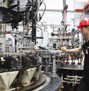 Xinghuo Glass's modern, large-capacity automated glass production line (Strong Manufacturing Capacity) factory: Real-life images of heavy machinery and professional quality control operations. This is a close-up, medium-range realistic photograph taken inside a modern industrial plant, showcasing Xinghuo Glass's automated manufacturing capabilities and the details of its glass production line. On the left and center of the image stands a massive, complex, modern, multi-station rotary glass pressing or blowing machine. The steel frame is supported by numerous numbered, off-white conical mold bases, while above it are a dense network of metal pneumatic and hydraulic transmission pipes, mechanical rocker arms, spring suspension components, and a complex web of black control cables. The entire large machine exudes a robust industrial metallic feel. In the background, the spacious, tall white steel structure of the factory building and other large industrial furnaces are faintly visible. On the right side of the image, a young, capable Asian male technician stands sideways beside the machine, wearing a bright red standard construction helmet and black short-sleeved work clothes. His right hand rests on the silver metal control handle in the center of the machine, his expression focused and his gaze intently fixed on the operation of the automated turntable. The entire photo is realistically lit and without any artificial embellishment, perfectly integrating the cutting-edge, modern heavy-duty manufacturing equipment for everyday glass with rigorous and safe front-line production operations, fully demonstrating the strong foundation of this large foreign trade factory in terms of mass production and fulfilling large-volume orders.