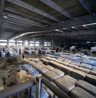 Real-world images of ultra-large capacity industrial warehousing and multinational large-volume digital drop-and-hook logistics bases (Reliable Wholesale Supply Chain) – mature supply chain fulfillment yards of major manufacturers. This is a hardcore, realistic photograph taken from a high-angle, overhead perspective, designed to showcase the massive industrial warehousing and reliable wholesale supply chain of a major B2B export manufacturer. The main subject of the image is an extremely spacious, tall, and seemingly bottomless modern steel-structured logistics warehouse hall. The high ceiling is supported by dense gray I-beams and metal trusses, with rows of industrial spotlights hanging in the depths, projecting bright white ribbons of light. On the entire open warehouse floor, thousands upon thousands of massive quantities of goods are densely and meticulously stacked, arranged in dozens of long matrix lines: the goods in the foreground and midground on the right are tightly wrapped in thick, light brown moisture-proof kraft paper and transparent bubble wrap, stacked into rows of half-meter-high rectangular stacks; the goods in the background on the left are wrapped in blue and white striped rainproof high-strength plastic protective film, also arranged in rows and matrices extending endlessly towards the light-filled door at the far end of the warehouse. On the left, in a spacious, sturdy gray concrete walkway marked with signs of work and dampness, a warehouse worker in dark clothing moves almost microscopically between the cargo areas, emphasizing the sheer size of the warehouse space. A section of metal railing juts out at an angle in the foreground. The photograph, with its strong contrast of light and shadow, profound sense of depth, and impressive scale of goods, vividly portrays the efficient logistics of a modern large-scale factory, its massive inventory, and its robust multinational supply chain ensuring stable, high-volume delivery.