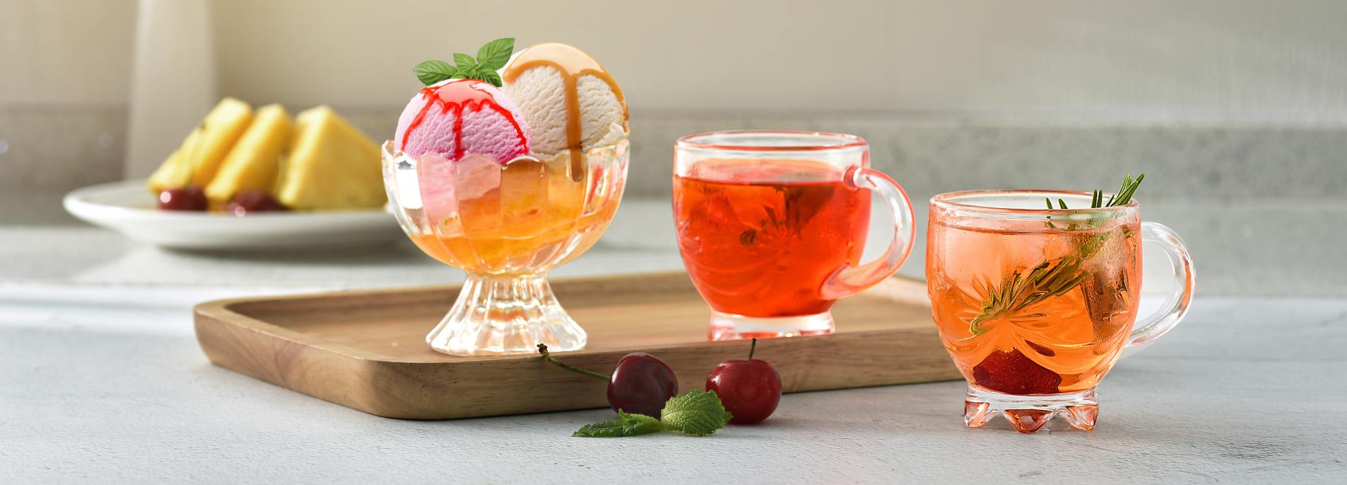 A real-life marketing photo taken in an ultra-wide frame, in a bright and airy indoor afternoon tea setting on a summer afternoon, aimed at promoting Xinghuo Glass's handle-carved embossed glass mugs and stemmed dessert bowls (Mug cups). On a light gray m