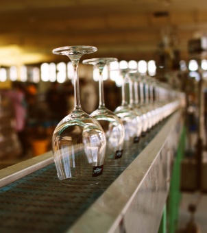 This is a marketing photograph taken with a medium-close-up shot inside a busy and large-scale modern glass manufacturing workshop, intended to promote Xinghuo Glass's automated crystal goblet production line. On a straight, automated industrial conveyor belt with a metal frame, a row of highly transparent, lead-free crystal goblets are neatly and uniformly inverted on the green checkered conveyor belt. The focus is on the first goblet in the foreground, its large, round rim facing downwards, its pure, transparent walls clearly reflecting the light and shadow of the factory. A small, fine, dark rectangular label is affixed to the glass; its slender, one-piece stem points straight into the air, connecting to a smooth, rounded base. The line of sight extends infinitely to the right and rear of the conveyor belt, where dozens of identical goblets at the same production stage are neatly arranged in a straight line, gradually shrinking and blurring as the depth of field increases, creating a visually striking sense of industrial order. The conveyor belt is painted green on the side. In the blurred background to the left rear of the image, the tall, warm-toned factory buildings, scattered machinery, and the figure of a quality inspector in work clothes are vaguely visible. The entire photo is naturally lit and has a strong sense of depth, perfectly combining the exquisite craftsmanship of crystal goblets with large-scale industrial assembly line manufacturing, fully demonstrating the professional supply chain strength of a major export-grade household and commercial glassware manufacturer.