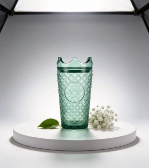 The Xinghuo Glass, a dark green crown-capped fish-scale patterned glass, is displayed on a white circular pedestal, surrounded by white flowers and green leaves.